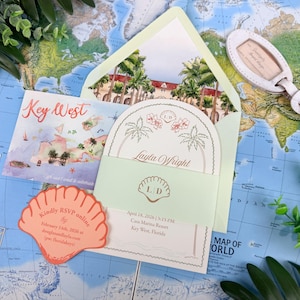 May include: A wedding invitation suite featuring a pale green envelope with a watercolor illustration of a resort, a card with the name Layla Wright, a shell-shaped RSVP card, and a luggage tag, all displayed on a world map background.