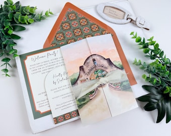 Può includere: Un set di partecipazioni di nozze con un biglietto a libro illustrato ad acquerello del villaggio artigianale di Tlaquepaque, legato con un nastro color crema. Include un biglietto di benvenuto e una scheda RSVP con bordi a motivi arancioni.