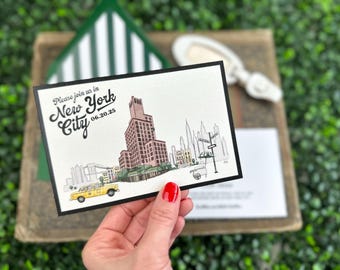 May include: A rectangular wedding invitation card with a black border featuring a hand-drawn illustration of the New York City skyline, a yellow taxi, and text that reads Please join us in New York City 06.20.25 in black script and print.