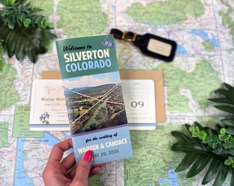 Può includere: Un invito di nozze con una fotografia paesaggistica di Silverton, Colorado, con il testo Welcome to SILVERTON COLORADO for the wedding of WARREN & CANDACE JUNE 20, 2026, legato con dello spago, posizionato sopra una mappa con un'etichetta.