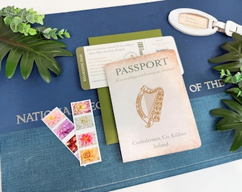 Può includere: Un set di inviti di nozze composto da un libretto in stile passaporto con un'illustrazione di un'arpa dorata, una carta d'imbarco verde, due strisce di francobolli floreali e un'etichetta per bagagli bianca su una superficie blu.