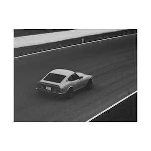 Könnte beinhalten: Ein Schwarzweißfoto eines silbernen Oldtimer-Sportwagens, der auf einer Rennstrecke fährt.