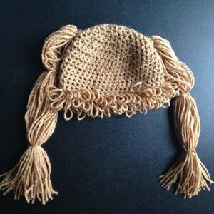 May include: A crocheted hat with long, brown yarn strands resembling pigtails. The hat is beige and has a textured, crocheted design.