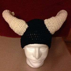 May include: A black and white crocheted hat with two long white crocheted horns. The hat is on a white mannequin head.