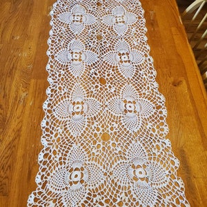 May include: White crocheted table runner with a delicate floral pattern. The runner is made of a fine thread and has a scalloped edge.
