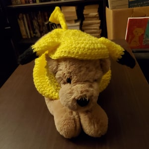 May include: A plush toy dog wearing a yellow crocheted banana costume. The dog is lying on a wooden surface.