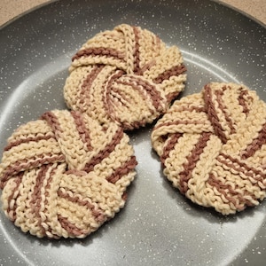 May include: Three circular, hand-knitted scrubbers featuring a spiral pattern in alternating cream and dusty rose yarn. Each scrubber has a textured, knotted construction with a dense, woven appearance, arranged together on a gray speckled surface.