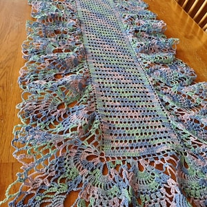 May include: A crocheted table runner with a lacy border in shades of blue, green, and pink. The runner is made with a delicate stitch pattern and has a fringed edge.