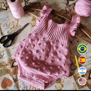 Puede incluir: Un pelele de bebé de ganchillo rosa con un patrón texturizado de punto bobble. El pelele tiene tirantes con volantes y un cierre de botón.
