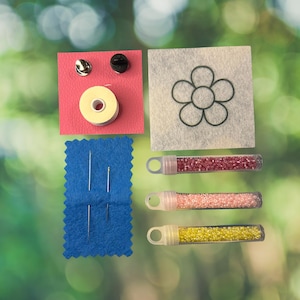May include: A craft kit containing a pink square with two metal pin backs and a spool of white thread, a white felt square with a black flower outline, a blue felt rectangle with four sewing needles, and three clear tubes filled with pink, peach, and yellow beads.