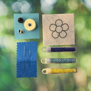 May include: A craft kit containing a light blue textured square with a spool of thread and two metal pin backs, a white felt square with a black flower outline, a blue felt rectangle with two needles, and three tubes of blue, teal, and yellow beads.