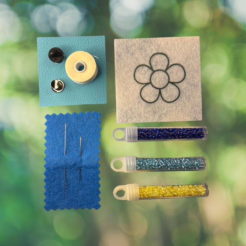 May include: A craft kit containing a light blue textured square with a spool of thread and two metal pin backs, a white felt square with a black flower outline, a blue felt rectangle with two needles, and three tubes of blue, teal, and yellow beads.