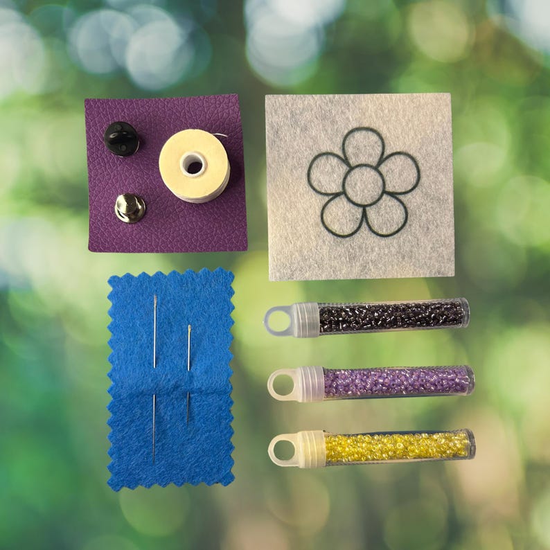 May include: A craft kit containing a purple textured square with two metal pin backs and a small spool of thread, a white felt square with a black flower outline, a blue felt rectangle with four needles, and three clear tubes filled with beads.