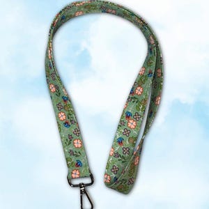 Op de afbeelding: Een groene bloemenlanyard met een metalen sluiting. De lanyard heeft een herhalend patroon van roze, blauwe en witte bloemen met groene bladeren op een groene achtergrond. De sluiting is zilver.