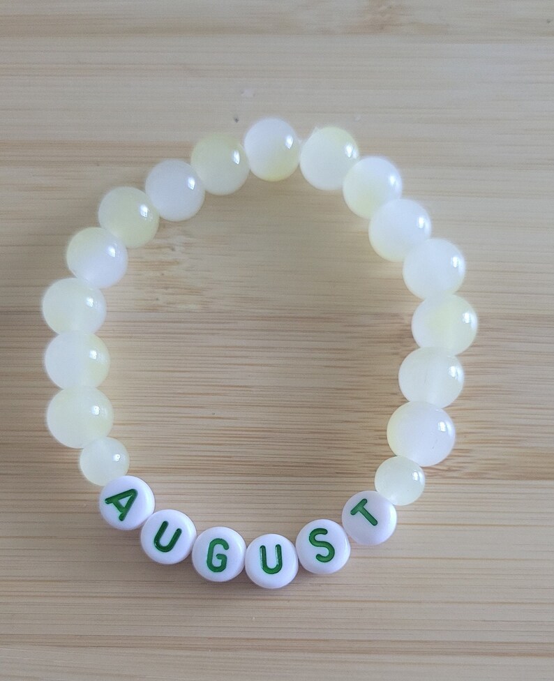 May include: A beaded bracelet with pale yellow and white round beads. The bracelet features white letter beads spelling "AUGUST" in green letters. The bracelet is displayed on a light brown wooden surface.