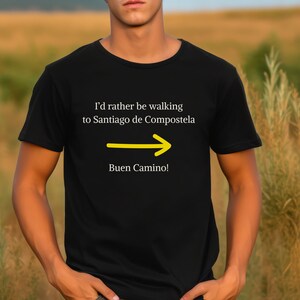 May include: A black t-shirt with white text that reads "I'd rather be walking to Santiago de Compostela" with a yellow arrow pointing to the right and the text "Buen Camino!" below the arrow.
