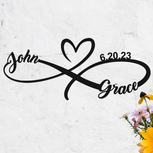 Può includere: Arte da parete in metallo nero con i nomi "John" e "Grace" intrecciati in un simbolo infinito con un cuore e la data "6.20.23". Un vaso di fiori è nell'angolo in basso a destra.