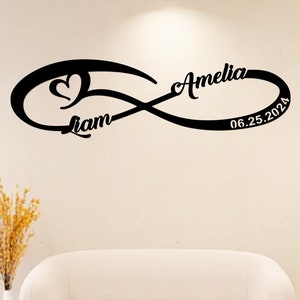 May include: Black metal infinity sign with a heart and the names "Liam" and "Amelia" in cursive script. The date "06.25.2024" is also included. The sign is mounted on a white wall.