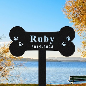 May include: Black metal dog bone-shaped memorial stake with the name "Ruby" and dates "2015-2024" in white. Paw prints are on each end. The stake is in a grassy area with a lake and trees in the background.