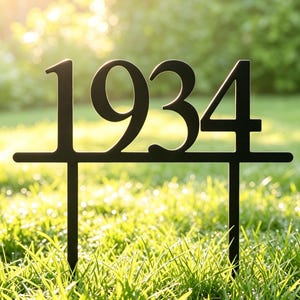 May include: A black metal garden stake featuring the numbers 1934 in a bold, sans-serif font. The numbers are connected by a horizontal bar and supported by two vertical prongs designed for insertion into the ground for outdoor display.