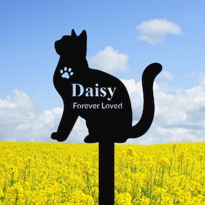 Könnte beinhalten: Schwarze Metallsilhouette einer Katze mit dem Namen Daisy und den Worten "Forever Loved" auf einem Pfahl. Der Pfahl steht in einem Feld mit gelben Blumen.