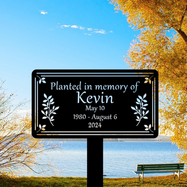 May include: Black metal memorial sign with white lettering. It reads "Planted in memory of Kevin May 10 1980 - August 6 2024". The sign has a leafy design.