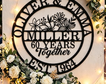 Könnte beinhalten: Schwarzes Metallschild mit den Namen "Oliver & Emma" und "Miller" in einem kreisförmigen Design. Das Schild erinnert an "60 JAHRE Together EST 1964" und hat florale Akzente. Weiße Rosen und Grün umgeben das Schild.