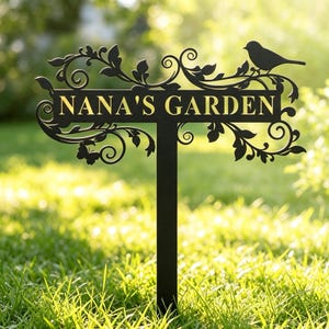 Puede incluir: Un estaca de jardín de metal negro con un letrero rectangular horizontal que muestra el texto NANA'S GARDEN en letras doradas. El letrero está decorado con volutas intrincadas, patrones de hojas y la silueta de un pájaro posado arriba.