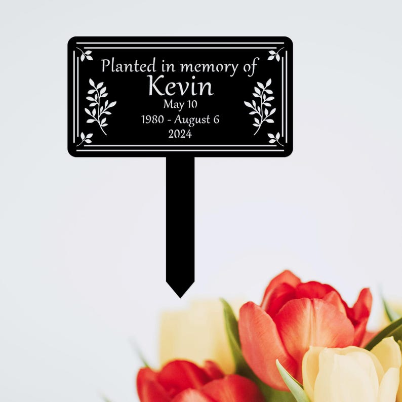 May include: Black metal garden marker with white text that reads "Planted in memory of Kevin May 10 1980 - August 6 2024".