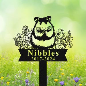 May include: Black metal garden stake featuring a hamster design with the name "Nibbles" and the years 2017-2024. The hamster is surrounded by floral cutouts. The stake is designed to be placed in a garden or yard.
