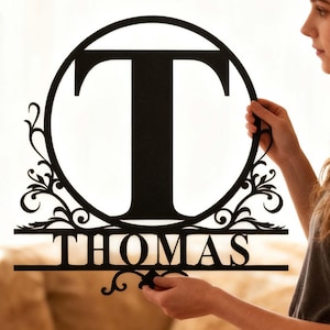 Pode incluir: Placa de monograma em metal preto com a letra "T" dentro de um círculo, com o nome "THOMAS" abaixo. O design inclui elementos decorativos. Uma pessoa está segurando a placa.