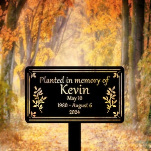 May include: Black metal memorial sign with gold lettering. The sign reads "Planted in memory of Kevin May 10 1980 - August 6 2024". The sign is surrounded by a gold leaf design.