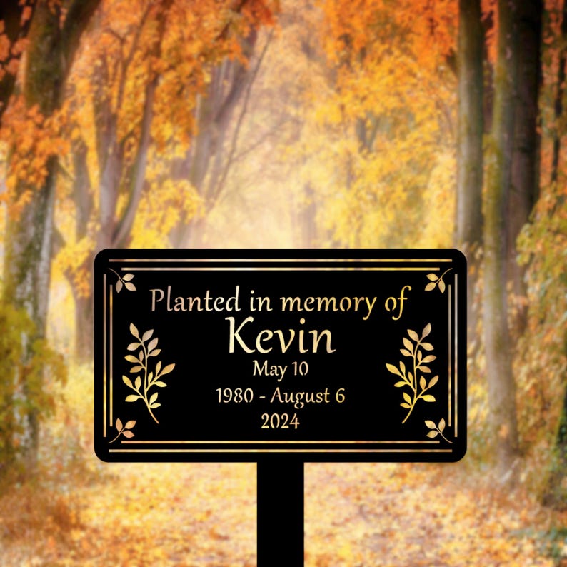May include: Black metal memorial sign with gold lettering. The sign reads "Planted in memory of Kevin May 10 1980 - August 6 2024". The sign is surrounded by a gold leaf design.
