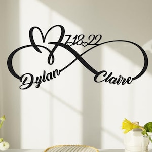 Pode incluir: Arte de parede em metal preto com um símbolo de infinito entrelaçado com um coração. Os nomes "Dylan" e "Claire" estão escritos no símbolo do infinito, com a data "7.18.22" acima. A obra de arte é exibida contra uma parede branca.