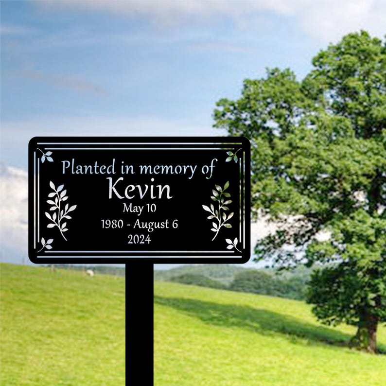 May include: Black metal memorial sign with the text "Planted in memory of Kevin May 10 1980 - August 6 2024" and two stylized leaf designs.
