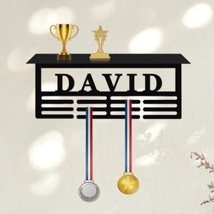 Może przedstawiać: Czarny metalowy wyświetlacz na trofea i medale z wyciętym imieniem "DAVID". Zawiera złote trofeum, trofeum w kształcie gwiazdy i dwa medale z czerwonymi, białymi i niebieskimi wstążkami. Dekoracyjny element do eksponowania osiągnięć.