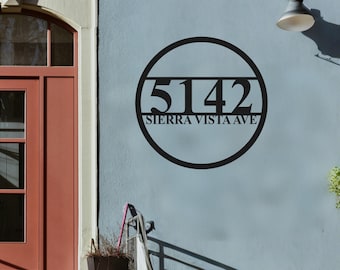 Puede incluir: Letrero de dirección de metal negro con los números "5142" y el texto "SIERRA VISTA AVE" dentro de un marco circular. El letrero está montado en una pared azul claro, cerca de una puerta roja y una lámpara de pared.