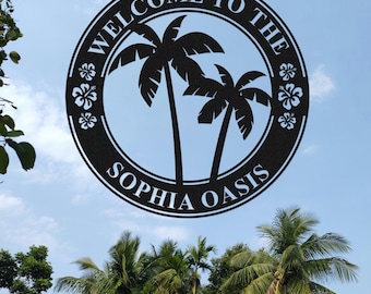 Könnte beinhalten: Schwarzes Metallschild mit den Worten "WELCOME TO THE SOPHIA OASIS" in einem kreisförmigen Design, mit Palmen und floralen Akzenten. Das Schild befindet sich vor einem blauen Himmel mit Palmen im Hintergrund.