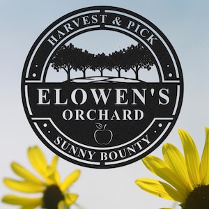 Puede incluir: Un letrero de metal negro con las palabras "HARVEST & PICK" y "ELOWEN'S ORCHARD SUNNY BOUNTY" en un diseño circular. El letrero presenta una silueta de árboles y una manzana. Girasoles amarillos en primer plano.