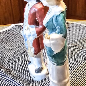 以下が含まれることがあります： 植民地時代の服装をした人々の磁器製フィギュア2体。左側のフィギュアは赤いジャケットと白いズボンを着用しています。右側のフィギュアは緑色のドレスと白いスカートを着用しています。両方のフィギュアは黒と白のパターンが施された表面に立っています。