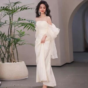 May include: A woman wearing a white off-the-shoulder gown with a flowing skirt and puffy sleeves. The gown is made of a soft, silky fabric and has a fitted bodice.