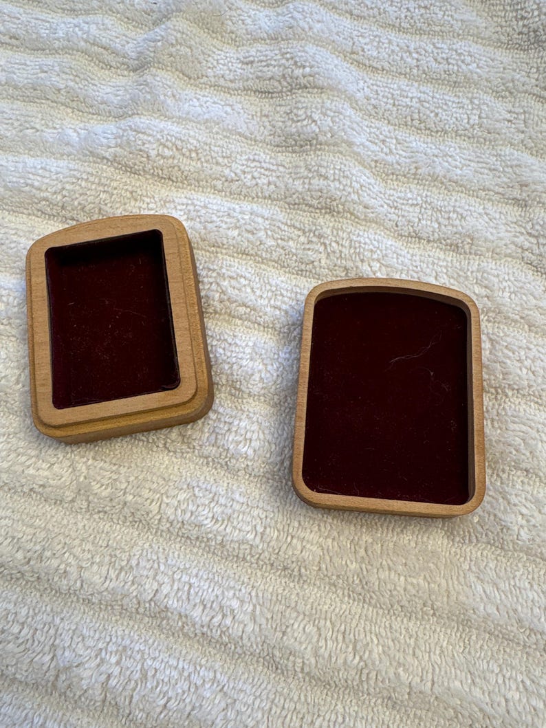 K&ouml;nnte beinhalten: Zwei kleine, rechteckige Holzkisten mit dunkelrotem Samtfutter. Die Kisten sind ge&ouml;ffnet und geben den Innenraum frei. Das Holz hat eine hellbraune Farbe. Die Kisten befinden sich auf einer wei&szlig;en, strukturierten Oberfl&auml;che.