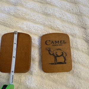 K&ouml;nnte beinhalten: Zwei rechteckige Untersetzer aus Holz. Einer ist schlicht, der andere zeigt das Wort "CAMEL" &uuml;ber einer Kamelillustration und dem Text "SINCE 1913". Die Untersetzer sind etwa 7,6 cm lang.