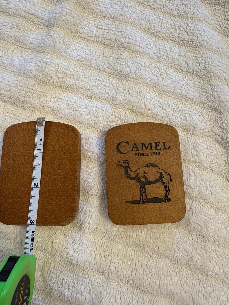 K&ouml;nnte beinhalten: Zwei rechteckige Untersetzer aus Holz. Einer ist schlicht, der andere zeigt das Wort "CAMEL" &uuml;ber einer Kamelillustration und dem Text "SINCE 1913". Die Untersetzer sind etwa 7,6 cm lang.