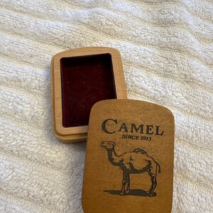K&ouml;nnte beinhalten: Rechteckige Holzbox mit Scharnierdeckel. Der Deckel zeigt das Wort "Camel" &uuml;ber einer Kamelillustration und dem Text "SINCE 1913". Der Innenraum der Box ist mit bordeauxfarbenem Samt ausgekleidet.
