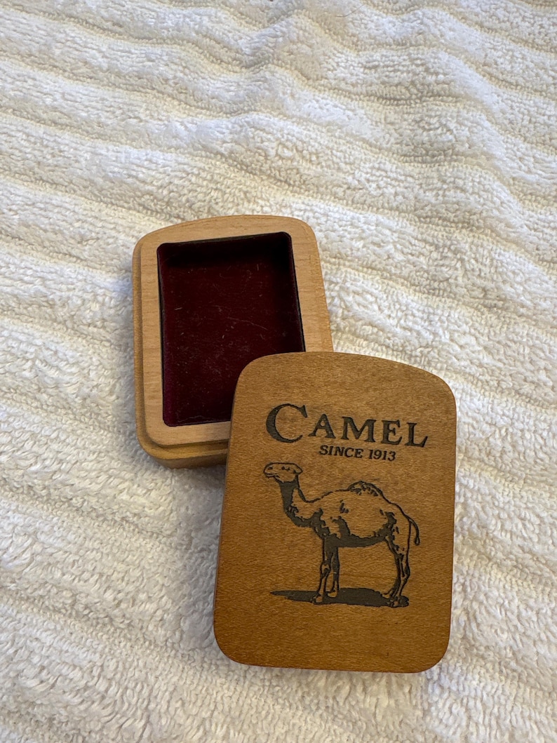 K&ouml;nnte beinhalten: Rechteckige Holzbox mit Scharnierdeckel. Der Deckel zeigt das Wort "Camel" &uuml;ber einer Kamelillustration und dem Text "SINCE 1913". Der Innenraum der Box ist mit bordeauxfarbenem Samt ausgekleidet.