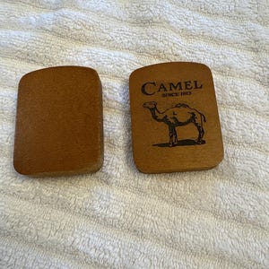 K&ouml;nnte beinhalten: Zwei Holzartikel der Marke Camel. Einer ist schlicht, der andere zeigt das Camel-Logo mit einer Kamelsilhouette und dem Text "CAMEL SINCE 1913". Beide Artikel sind rechteckig mit abgerundeten Ecken.