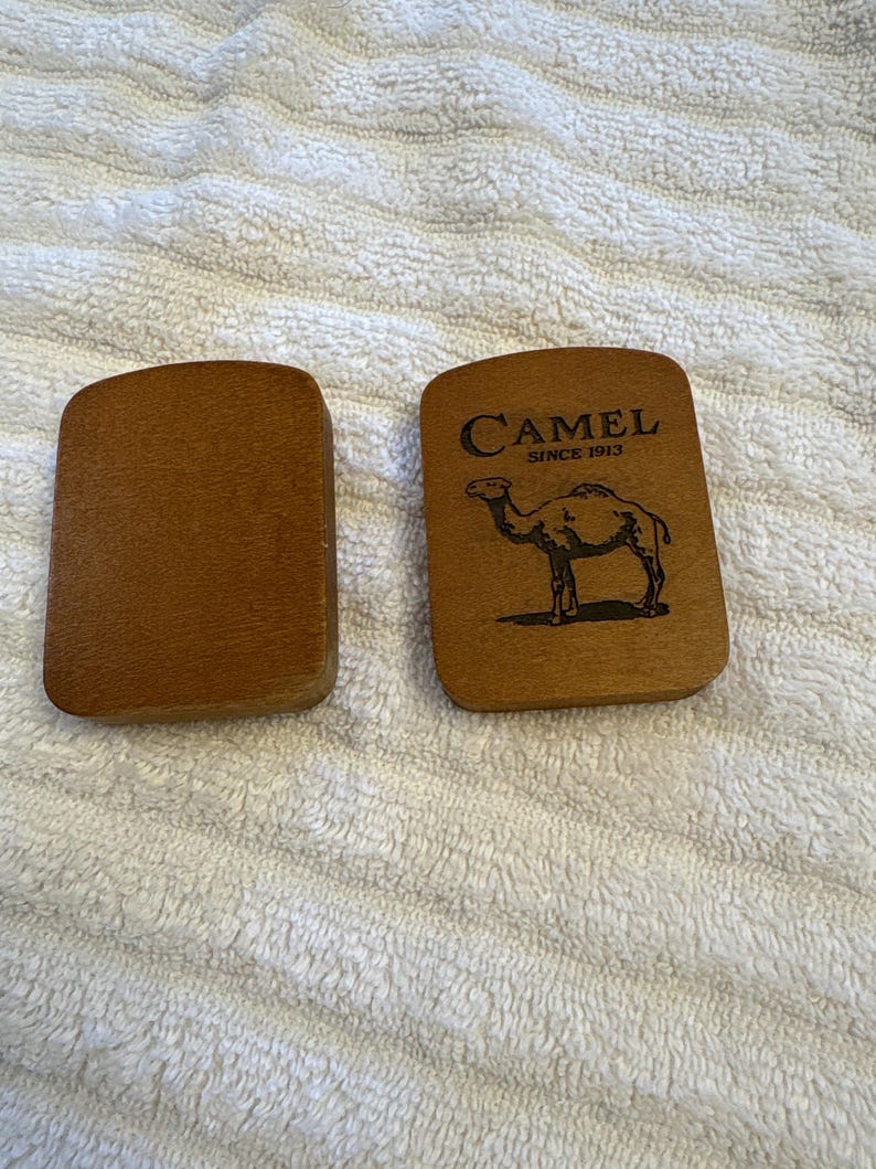K&ouml;nnte beinhalten: Zwei Holzartikel der Marke Camel. Einer ist schlicht, der andere zeigt das Camel-Logo mit einer Kamelsilhouette und dem Text "CAMEL SINCE 1913". Beide Artikel sind rechteckig mit abgerundeten Ecken.
