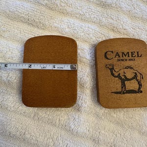K&ouml;nnte beinhalten: Zwei Untersetzer aus Holz. Einer wird mit einem Ma&szlig;band gemessen und ist etwa 7,6 cm lang. Der andere Untersetzer tr&auml;gt das Wort "Camel" und eine Kamelillustration. Die Untersetzer sind braun.