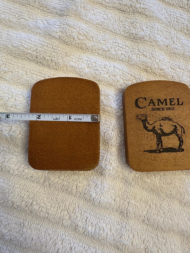 K&ouml;nnte beinhalten: Zwei Untersetzer aus Holz. Einer wird mit einem Ma&szlig;band gemessen und ist etwa 7,6 cm lang. Der andere Untersetzer tr&auml;gt das Wort "Camel" und eine Kamelillustration. Die Untersetzer sind braun.